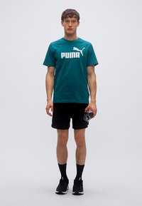 Μπλούζα από βαμβάκι σε τιρκουάζ με τολμηρό λευκό λογότυπο "PUMA", συνδυασμένη με μαύρο σορτς και αθλητικά παπούτσια. Το άτομο κρατά ένα διαφανές μπουκάλι νερού.