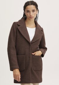 Manteau en laine marron avec une finition texturée, doté d'un large col, de deux poches avant et d'une coupe droite, associé à un haut blanc.