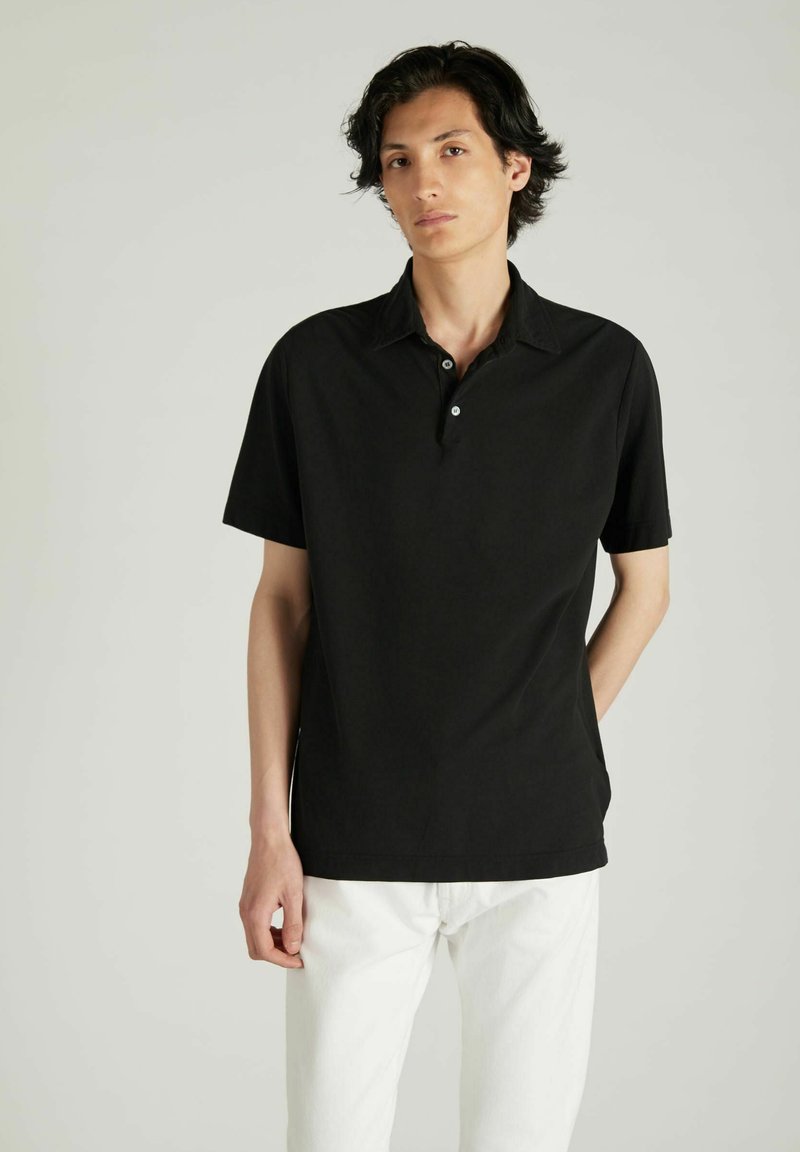Polo negro de manga corta con una tapeta de botones, hecho de una tela suave. El modelo lo lleva con pantalones blancos sobre un fondo neutral.