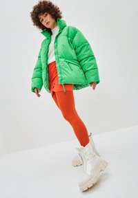 Chaqueta de plumas verde brillo, suéter blanco, falda mini naranja y leggings naranjas. Botas blancas gruesas con textura acolchada.
