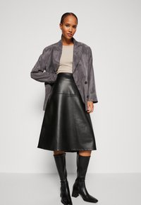 Siv suede blazer preko bež majice, uparen s crnom kožnom suknjom do koljena i crnim čizmama na petu, stvarajući modernu siluetu.