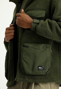 Jasnozielona polarowa kurtka z teksturowanej tkaniny, z kieszeniami na zamek i płaską kieszenią. Posiada elastyczne mankiety oraz zamek błyskawiczny.