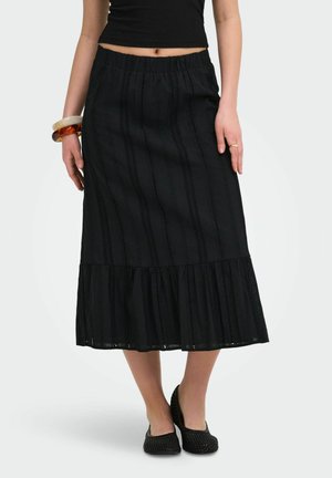 Femme portant une jupe midi noire avec des bandes de dentelle verticales et un ourlet froncé, associée à des ballerines tissées noires et des bracelets larges.