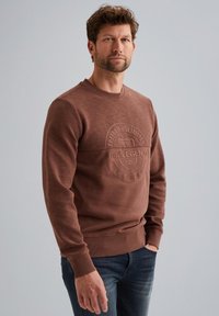 Smeđa dukserica s teksturiranom prednjom stranom, s urezanim logotipom i rebrastim manžetama. U kombinaciji s tamnoplavim trapericama. Lagana tkanina.