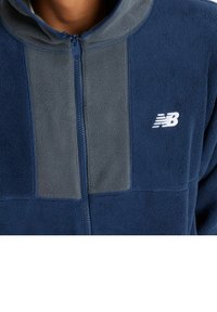 Mörkblå fleecetröja med en grå panel, hel dragkedja och en vit logotyp på bröstet. Mjuk textur, avslappnad passform och ribbad krage.