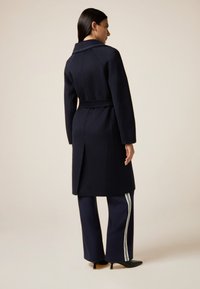 Cappotto di lana navy con vita regolabile, caratterizzato da un colletto a revers e uno spacco sul retro, abbinato a pantaloni navy con strisce bianche sui lati.