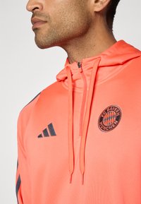 Oransje treningsgenser med glidelås i kragen, med mørkegrå detaljer og FC Bayern München-logo på brystet. Glatt tekstur, lett stoff.