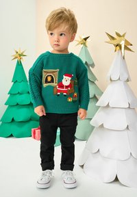 Kleinkind mit blonden Haaren, das einen türkisfarbenen Weihnachtspullover mit einem Weihnachtsmann und Geschenken trägt. Es steht neben weißen und grünen Papier-Weihnachtsbäumen, die mit goldenen Sternen geschmückt sind.
