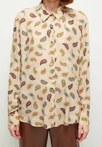 Blusa de manga larga y botones en tela beige con un estampado de paisley en rojo, verde y amarillo. Presenta un cuello clásico y un dobladillo redondeado.