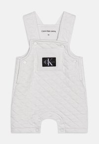 Pikowane białe kombinezon z regulowanymi ramiączkami, kieszenią z przodu i zatrzaskami na dole. Posiada czarną naszywkę z logo na kieszeni.