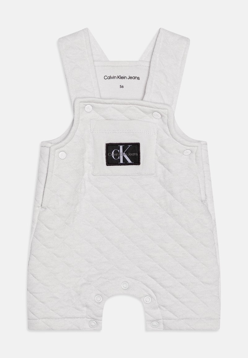 Pikowane białe kombinezon z regulowanymi ramiączkami, kieszenią z przodu i zatrzaskami na dole. Posiada czarną naszywkę z logo na kieszeni.