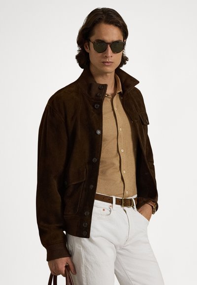Brązowa zamszowa kurtka z kołnierzem zapinanym na guziki, przednimi kieszeniami i ściągaczami przy mankietach, zestawiona z beżową, teksturowaną koszulą oraz białymi spodniami.