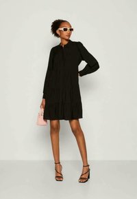 Svart långärmad klänning med en flerlagrig design, lättviktsmaterial och knäppning. Matchad med svarta remsandaler och en rosa handväska.