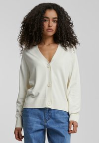 Jeune femme aux cheveux bouclés portant un cardigan blanc à boutons et un jean bleu sur un fond gris uni.