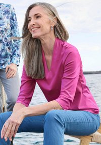 Top fucsia con scollatura a V e maniche tre quarti, realizzato in tessuto liscio. Indossato con jeans blu, seduto vicino all'acqua. Capelli grigi lunghi e orecchini visibili.
