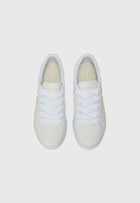 Hvite joggesko med glatt skinnoverdel, flate hvite snører, beige detaljer og en teksturert såle. Lacoste-logo på sidene.