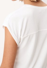 Detailní záběr na horní část zad a rameno osoby, která má na sobě bílou košili s krátkým rukávem a vyhrnutými manžetami, a má světlou pleť.