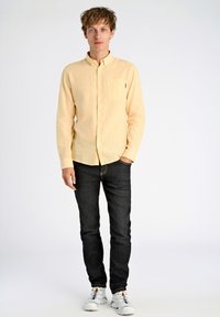 Lichtgele lange mouw knoopshirt met een zak, gecombineerd met donkere jeans en witte sportschoenen met kleurrijke accenten.