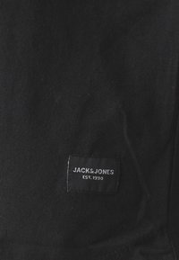 Sort bomuldsstof med et vævet label, der har "JACK & JONES" og "EST. 1990" i hvid tekst, placeret i nederste venstre hjørne.