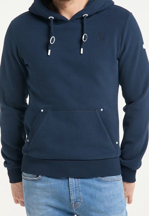Mann trägt marineblauen Kapuzenpullover mit Fronttasche, Kordelzug und dezenter Anker-Logos auf der Brust, kombiniert mit hellblauen Jeans.