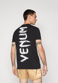 Črna bombažna majica s kratkimi rokavi, ki ima na hrbtu velik, razpokan bel napis "VENUM" v pokončni poziciji. Oblika ovratnika je krožna, model pa nosi bež kratke hlače.