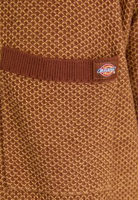 Ruda tekstūruota megztinė medžiaga su deimanto raštu. Yra juosmens juosta su briaunomis ir mažas Dickies logotipo ženklelis šone.