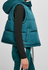 Tyrkysová péřová vesta s kapucí, s předním zipem, bočními kapsami a prošívanou strukturou. Kombinováno s tmavými dlouhými rukávy a tepláky.