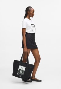 Fekete vászontáska, napszemüveges férfi fényképnyomattal, két erős pánttal és sima felülettel. A modellen fehér póló és fekete szoknya van.