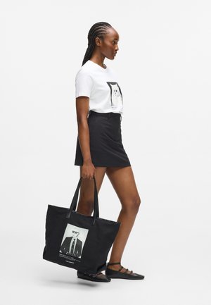 Geantă tote neagră cu un imprimeu foto al unui bărbat în ochelari de soare, două mânere rezistente și o textură netedă. Modelul poartă un tricou alb și o fustă neagră.