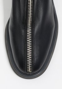 Botín de cuero negro con un prominente zipper plateado en la parte delantera, punta redondeada y suela de goma texturizada.