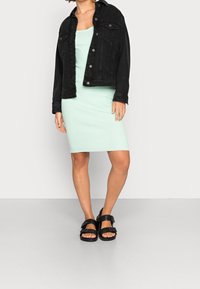 Chaqueta de mezclilla negra sobre un vestido midi de canalé verde menta. El conjunto incluye sandalias negras gruesas con hebillas en la parte delantera.