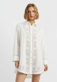 Camicia bianca con bottoni realizzata in cotone, caratterizzata da ricami a taglio lungo la parte anteriore e le maniche, con un orlo posteriore leggermente allungato.