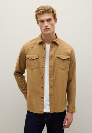 Camicia button-up tan realizzata in tessuto morbido, dotata di due tasche sul petto, bottoni a pressione e una vestibilità rilassata, indossata sopra una maglietta bianca.