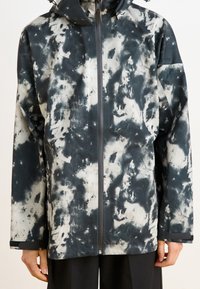 Widok z przodu wodoodpornej kurtki w czarno-białym wzorze tie-dye, z zamkiem błyskawicznym, kapturem i luźnym krojem. Gładka faktura i długie rękawy.