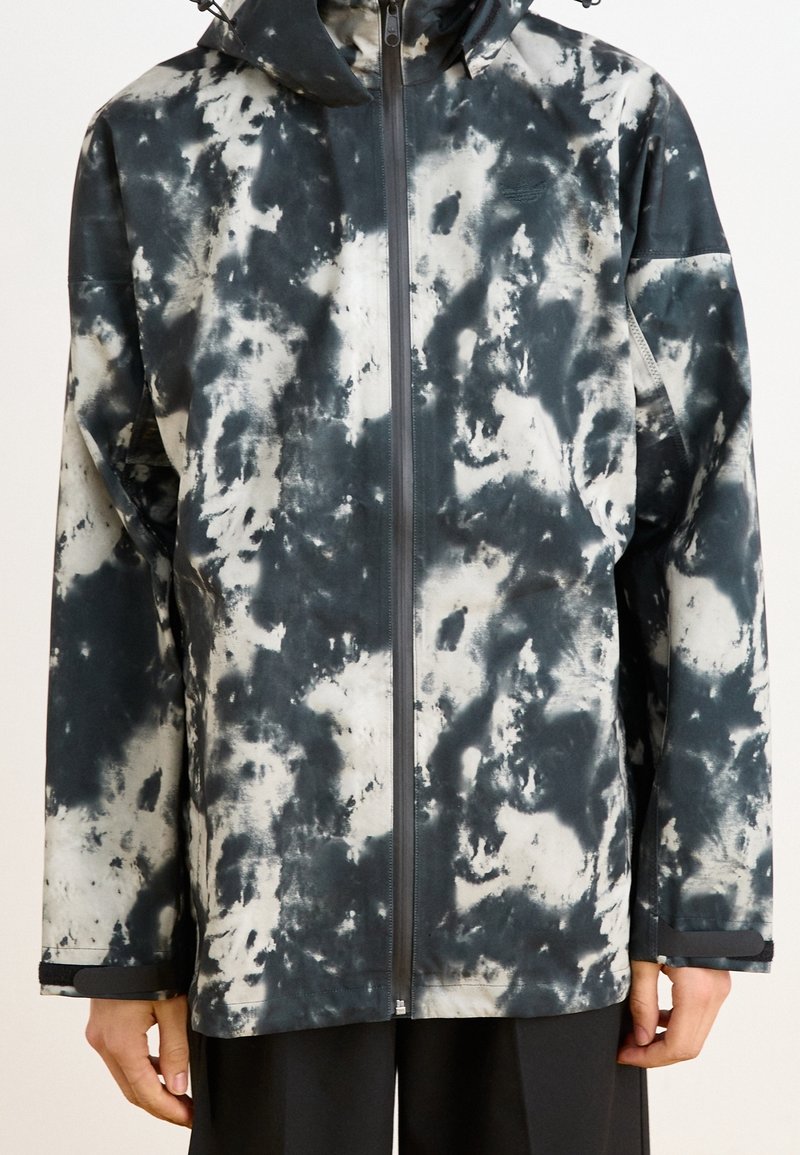 Widok z przodu wodoodpornej kurtki w czarno-białym wzorze tie-dye, z zamkiem błyskawicznym, kapturem i luźnym krojem. Gładka faktura i długie rękawy.