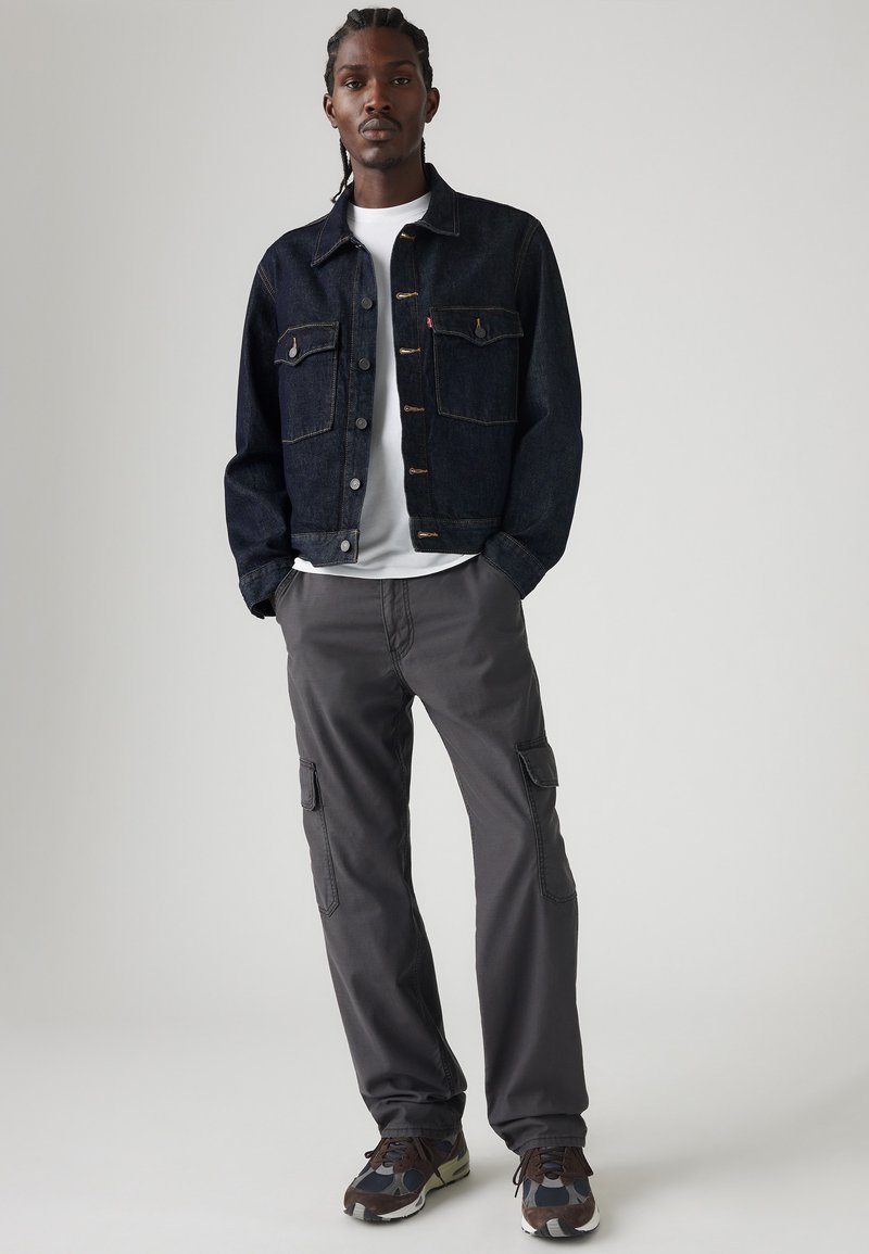 Chaqueta de mezclilla oscura con dos bolsillos en el pecho con botón, sobre una camiseta blanca. Pantalones cargo grises con bolsillos laterales y suelas texturizadas en múltiples colores.