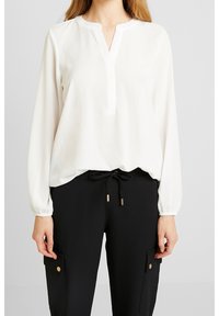 Donna che indossa una blusa bianca a maniche lunghe con colletto a fessura e pantaloni neri con coulisse e tasche laterali con bottone.