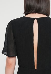 Svart kjole med teksturert stoff, korte gjennomsiktige ermer og en nøkkelhullåpning bak med en liten glidelås.