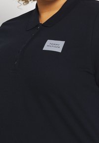 Námořnická modrá polo košile vyrobená z texturované látky, s tlačným límečkem a obdélníkovou nášivkou s nápisem "TOMMY HILFIGER" v bílé barvě.