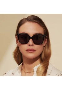Schwarze Cat-Eye-Sonnenbrille mit dunklen Gläsern, glänzender Oberfläche und dezentem Branding in der oberen Ecke; getragen von einer Person mit schulterlangem Haar.