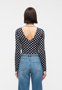 Γυναίκα με κοντά καστανά μαλλιά που φοράει μαύρο τοπ με μακριά μανίκια και λευκές πουά, με βαθύ V στην πλάτη, σε συνδυασμό με ψηλόμεσα μπλε τζιν.