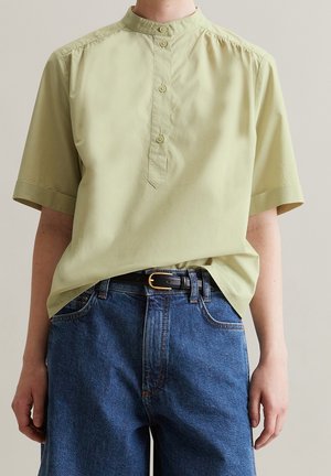 Persoon draagt een lichtgroen, kort mouwig doorgestikt hemd dat in een hoge taille blauwe jeans met een zwarte riem is gestopt.