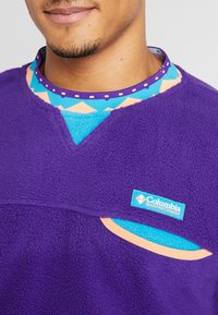 Pullover in fleece viola con colletto a motivo geometrico blu e corallo. Presenta una tasca e l'etichetta della Columbia Sportswear Company.