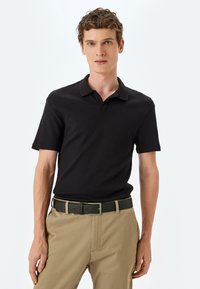 Camisa polo negra de canalé con cuello, mangas cortas, combinada con pantalones caqui y un cinturón negro. Diseño sencillo, líneas limpias.