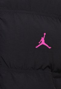 Svart stoff med en quiltet tekstur, med en fremtredende rosa brodert Jumpman-logo. Sømmene er synlige.