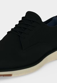 Baskets noires à lacets avec tige en tissu texturé et semelle beige sur fond blanc.