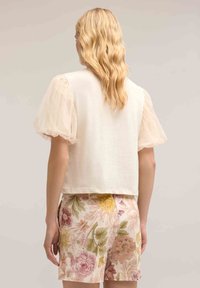 Kortærmet bluse med gennemsigtige, puffede ærmer; ensfarvet elfenbensfarvet krop. Sammen med blomstermønstrede shorts i pastelfarver.