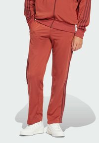 Pantalons de sport rouges avec des jambes fuselées, taille élastique et bandes latérales plus foncées. Associés à des baskets blanches, sur un fond clair.