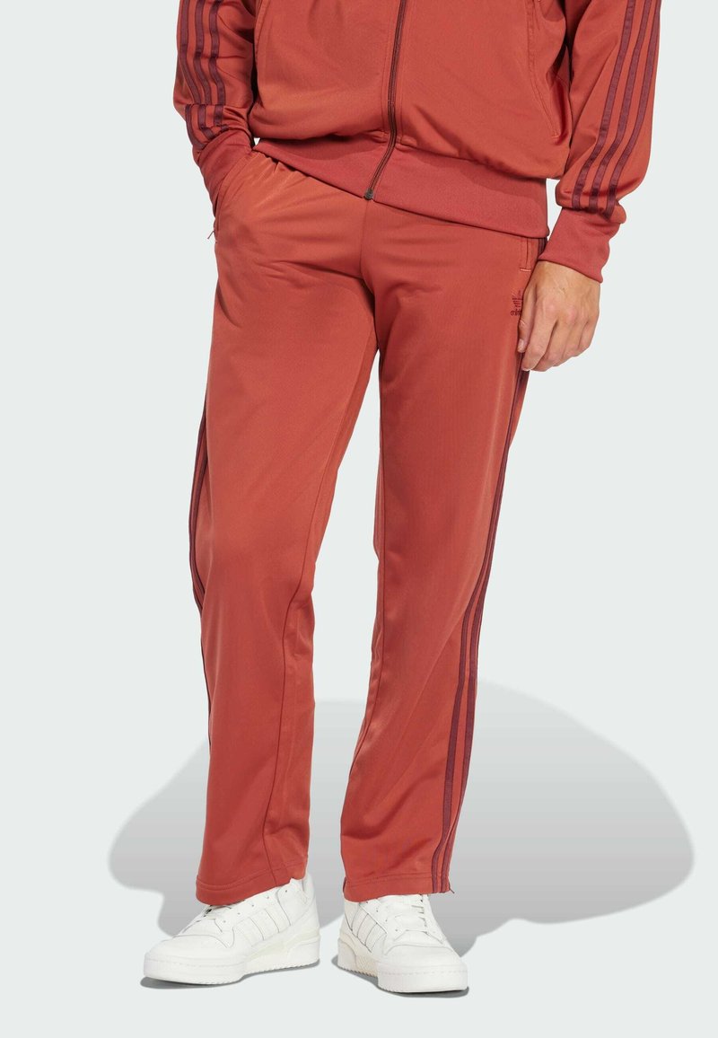 Pantalons de sport rouges avec des jambes fuselées, taille élastique et bandes latérales plus foncées. Associés à des baskets blanches, sur un fond clair.