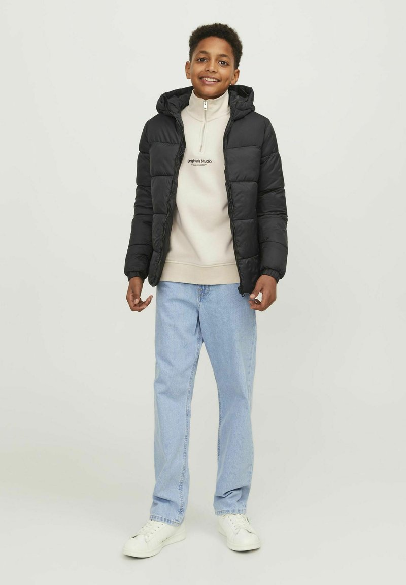 Doudoune noire avec capuche, portée sur un pull polaire zippé beige. Un jean bleu clair et des baskets blanches complètent la tenue.